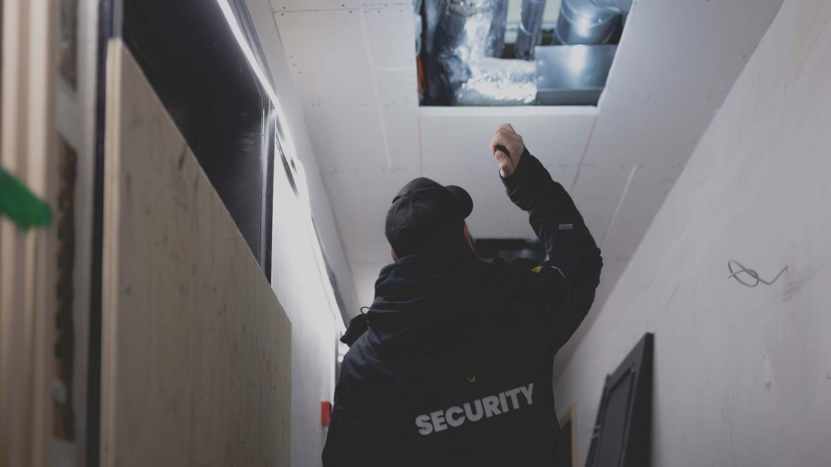 En vagt i sort jakke står på en gang og lyser op i et hul i loftet med sin lommelygte